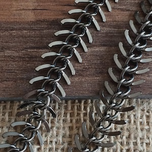 May include: A close-up of a black metal chain with a unique zig-zag pattern. The chain is made of small, interlocking links that create a textured and edgy look.