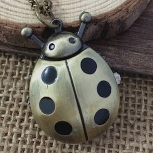 Puede incluir: Un collar de reloj de bolsillo de color dorado en forma de mariquita con manchas negras. El reloj tiene una esfera redonda y está unido a una cadena.