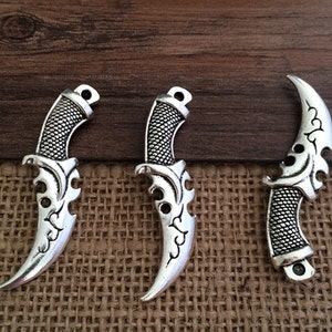 May include: Three silver-toned metal knife charms with intricate designs. The charms are shaped like daggers with a curved blade and a pointed tip. The charms have a textured surface and a loop at the top for attaching to a necklace or bracelet.