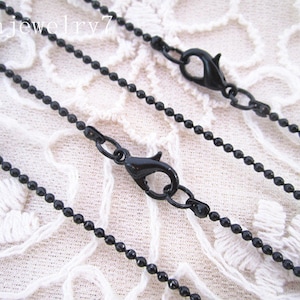 May include: Three black ball chain necklaces with lobster claw clasps. The chains are different lengths and are laid out on a white lace fabric.
