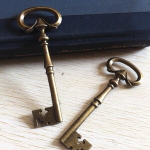 Könnte beinhalten: Zwei antike Skelettschlüssel mit verziertem Metall und einer angelaufenen goldenen Oberfläche. Die Schlüssel haben ein dekoratives Design und sind perfekt für Bastelprojekte.
