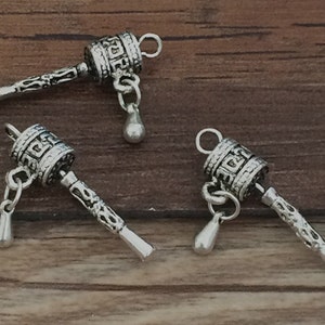 May include: Three silver prayer wheel charms with a decorative design. Each charm has a small silver drop hanging from it.