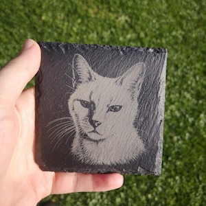 May include: A square black slate coaster featuring a laser-engraved portrait of a cat. The cat is depicted with detailed fur texture, pointed ears, and visible whiskers. The coaster has a rough, natural stone edge and a dark gray surface.