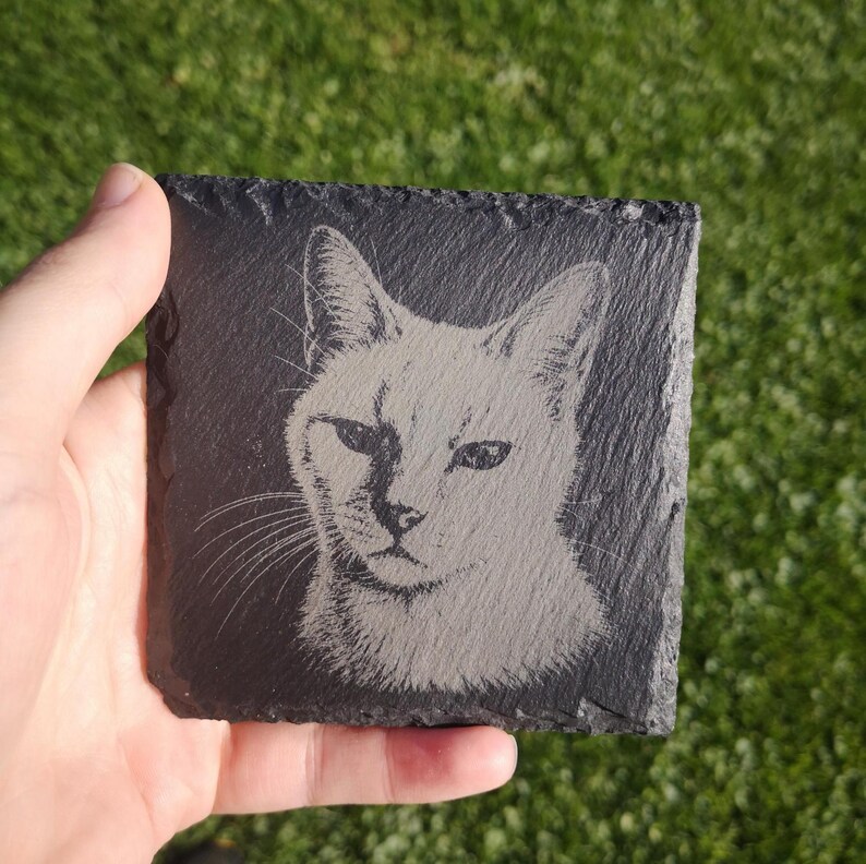 May include: A square black slate coaster featuring a laser-engraved portrait of a cat. The cat is depicted with detailed fur texture, pointed ears, and visible whiskers. The coaster has a rough, natural stone edge and a dark gray surface.