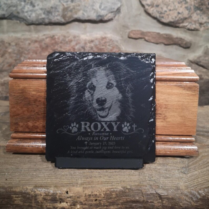 Może przedstawiać: Kwadratowa czarna tablica pamiątkowa z łupka z wygrawerowanym portretem psa. Tekst zawiera napis ROXY, Roxanne, Always in Our Hearts, January 27, 2023 oraz dedykację. Tablica ma surowe krawędzie i stoi na małej podstawce.