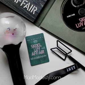 Może przedstawiać: Asortyment towarów, w tym pałeczka świetlna, płyta CD i karta fotograficzna, wszystkie z tekstem "SKOOL LUV AFFAIR". Przedmioty są ułożone na białej powierzchni, z czarną wstążką z napisem "ARMY".