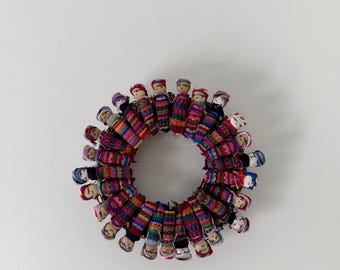 May include: A circular arrangement of numerous small, handcrafted dolls. Each doll is wrapped in vibrant, multi-colored striped fabric and features a simple, hand-painted face. The dolls are positioned side-by-side to form a continuous ring.