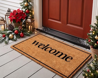 May include: A rectangular tan coir doormat with a black border and the word welcome printed in black script lettering. The mat is positioned on a porch in front of a red door, surrounded by holiday decorations including poinsettias and lights.