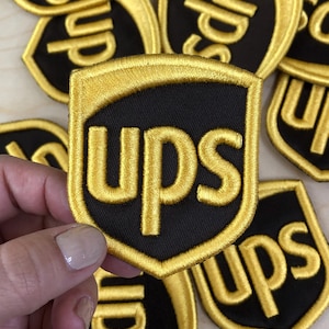 Pode incluir: Um emblema bordado em forma de escudo com fundo preto e as letras ups costuradas em fio dourado. O emblema tem uma borda dourada espessa e faz parte de uma coleção de emblemas idênticos com o mesmo design.