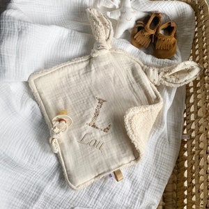 Peut inclure: Une couverture en mousseline blanche pour bébé avec un jouet en mousseline noué beige et blanc avec la lettre L et le nom Lou brodé dessus. Une étoile blanche avec un visage et des mocassins marrons sont également présents sur l'image.