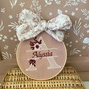 Könnte beinhalten: Ein rosa bestickter Reifen mit dem Namen "Alyssia" und einem floralen Design. Der Buchstabe "A" ist in Weiß gestickt. Eine florale Schleife in Weiß- und Burgundertönen ist oben am Reifen befestigt. Der Reifen wird auf einem Weidenkorb präsentiert.