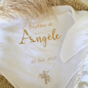 May include: A white muslin blanket featuring gold text that reads Baptême de Angèle, the date 10 Juin 2023, and a graphic of a cross with a dove. The letter A is stylized with floral elements. The fabric has a soft, crinkled texture throughout.