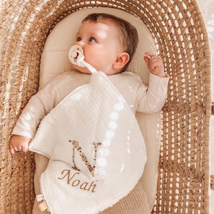 May include: A white muslin baby burp cloth with a floral embroidered letter N and the name Noah. The cloth is in a wicker basket with a baby wearing a white onesie and a pacifier.