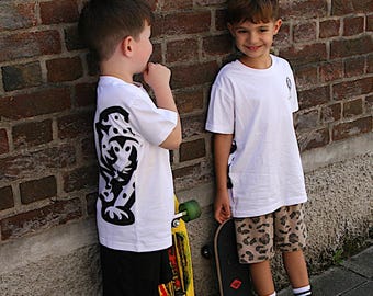 Könnte beinhalten: Zwei weiße T-Shirts mit schwarzen Grafikdrucken an den Seiten, eine schwarze Shorts, eine Shorts mit Leopardenmuster, weiße Socken mit schwarzen Streifen und Sportschuhe. Ein Kind hält ein gelbes Skateboard, das andere ein schwarzes.