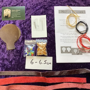 May include: A craft kit with various components on a purple surface. Includes a brown paper cutout, beads, leather strips, needles, and instructions. The kit also contains a business card and a label that reads "6-6.5cm".