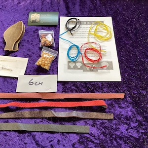 May include: A craft kit with various components on a purple surface. Includes leather strips, beads, string, and a paper with text. A card and a paper with "6cm" are also present. The kit appears to be for making jewellery or crafts.