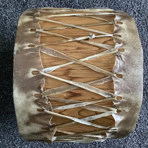 May include: A handmade drum featuring a wooden body and a stretched, light brown hide. The hide is laced across the wooden frame with leather strips. The drum's design suggests a focus on traditional craftsmanship.