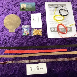 May include: A craft kit for a Native Echoes project, featuring instructions, beads, corn, leather strips, and a paper cutout. The kit includes a business card and a label that reads 7-8 cm. The components are arranged on a purple surface.