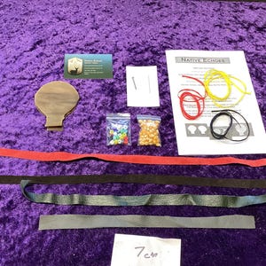 May include: A craft kit with a wooden shape, beads, leather strips, and cords. The kit includes a printed sheet titled "Native Echoes" and a business card. The items are arranged on a purple surface. The measurement "7 cm" is visible.