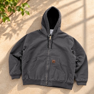 May include: A charcoal gray zip-up hooded sweatshirt featuring a black interior lining, a front kangaroo pocket with a small brown rectangular Carhartt brand patch, long sleeves with ribbed cuffs, and a metal zipper closure with a pull ring.