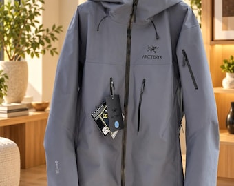 Könnte beinhalten: Eine schieferblaue Arc'teryx Kapuzenjacke mit durchgehendem schwarzem Frontreißverschluss, zwei vertikalen Brusttaschen und einer Ärmeltasche. Die Jacke zeigt ein Gore-Tex-Logo am Ärmel und ein hängendes Etikett mit der Größenangabe XL.