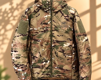 Op de afbeelding: Een jack met capuchon in een camouflagepatroon met bruine, groene en beige tinten. Het kledingstuk is voorzien van een ritssluiting over de gehele lengte, een klein Arc'teryx-logo op de borst en klittenband op beide mouwen.