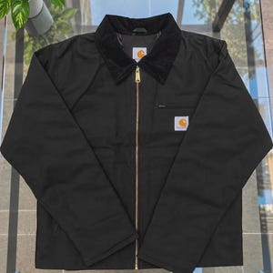May include: A black jacket featuring a corduroy collar, a full-length gold-tone metal zipper, and long sleeves. A white Carhartt brand label is stitched onto the left chest area, and a matching label is located inside the collar.