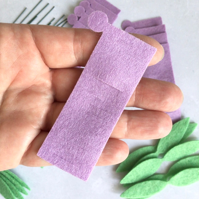 May include: A purple felt craft piece with a rectangular shape and a small square cut out in the center. The piece is held in a hand against a white background.