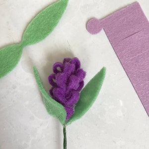 May include: A purple felt flower with green felt leaves attached to a brown stem. The flower is made of small, rolled felt balls.