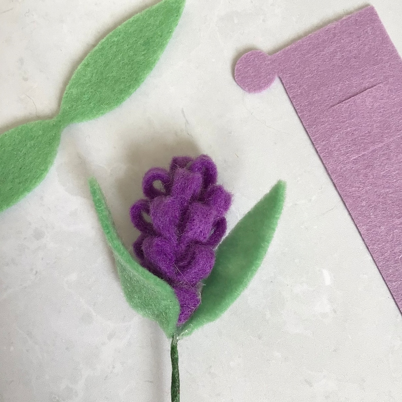 May include: A purple felt flower with green felt leaves attached to a brown stem. The flower is made of small, rolled felt balls.