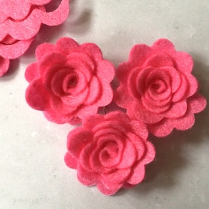 Puede incluir: Tres rosas de fieltro rosa con bordes festoneados. Las rosas están superpuestas para crear un efecto 3D.