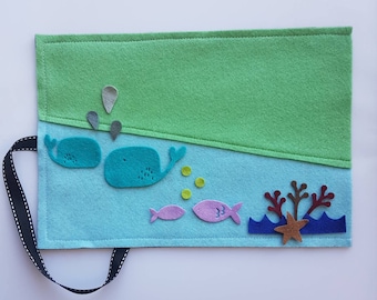 May include: A felt book with an ocean theme. The cover features a green sky and blue ocean with whales, fish, bubbles, and coral. A black and white ribbon is attached to the side. The book is designed for children.