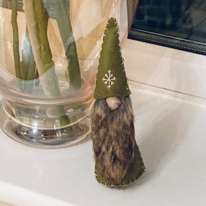 May include: A green felt gnome with a brown fur beard and a snowflake on its hat.