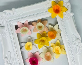 Può includere: Una collezione di fiori di narciso in feltro di vari colori, tra cui giallo, arancione, rosa e bianco. I fiori sono disposti in una cornice bianca.