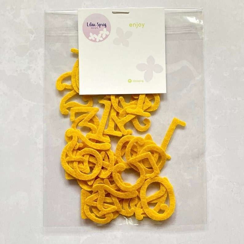 K&ouml;nnte beinhalten: Eine transparente Plastikt&uuml;te gef&uuml;llt mit gelben Filzbuchstaben, Zahlen von 0 bis 9. Die T&uuml;te hat ein wei&szlig;es Etikett mit dem Text "Lilac Sprig Designs" und "enjoy" darauf gedruckt.