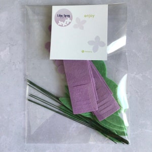 May include: A clear plastic bag containing a craft kit for making felt flowers. The kit includes purple felt flower petals, green felt leaves, and green wire stems. The bag also includes a card with the text "Lilac Sprig designs" and "enjoy".