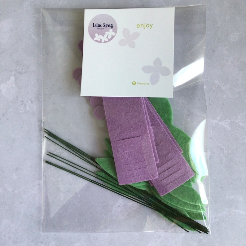 May include: A clear plastic bag containing a craft kit for making felt flowers. The kit includes purple felt flower petals, green felt leaves, and green wire stems. The bag also includes a card with the text "Lilac Sprig designs" and "enjoy".