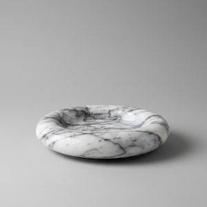 May include: A circular white marble tray featuring prominent grey veining throughout its polished surface. The tray has a thick, rounded rim and a shallow, concave center, showcasing the natural stone pattern in a smooth, glossy finish.