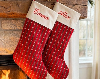 May include: Two Christmas stockings featuring a red fabric with vertical stripes and small floral patterns. Each stocking has a cream-coloured cuff with the names Emma and Alex embroidered in red script. The stockings hang from a wooden mantel.