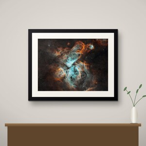 May include: A framed wall art print featuring a deep space nebula with vibrant swirls of bright cyan and burnt orange gas clouds set against a dark, star-filled background, enclosed in a simple black frame with a white mat border.