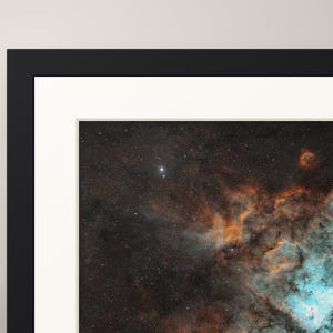May include: A framed wall art print featuring a deep space nebula with vibrant orange and turquoise gaseous clouds set against a dark, star-filled background. The artwork is enclosed in a thick black frame with a white mat border.