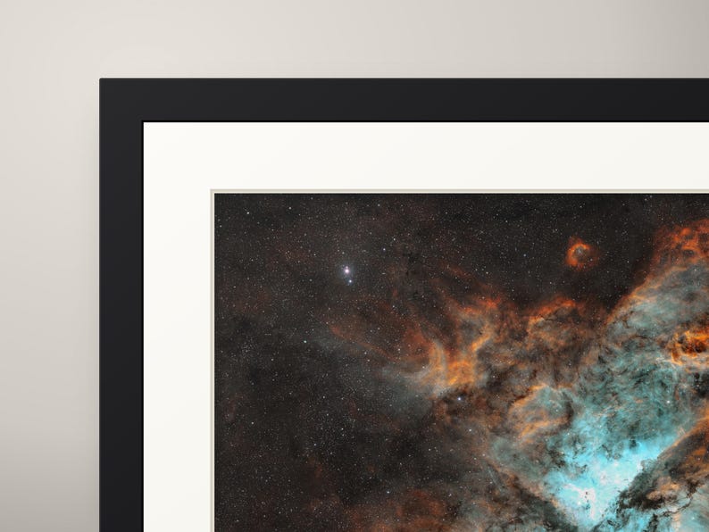 May include: A framed wall art print featuring a deep space nebula with vibrant orange and turquoise gaseous clouds set against a dark, star-filled background. The artwork is enclosed in a thick black frame with a white mat border.