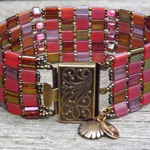 May include: A multi-strand bracelet made with pink, red, and brown glass beads. The bracelet has a brass clasp with a decorative floral design and a small seashell charm with the word "Ocean" on it.