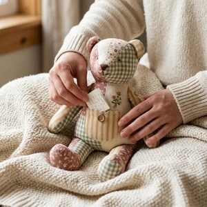 May include: A patchwork teddy bear constructed from various fabric patterns including green gingham, floral prints, and tan stripes. The bear features a small front pocket with a wooden button and holds a small white note with handwritten text.