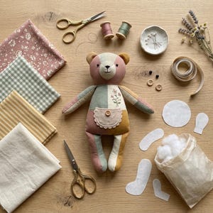 May include: A handmade patchwork teddy bear with a floral embroidered chest pocket, surrounded by sewing supplies including gold scissors, fabric swatches in floral, gingham, and striped patterns, thread spools, buttons, and a bag of stuffing.