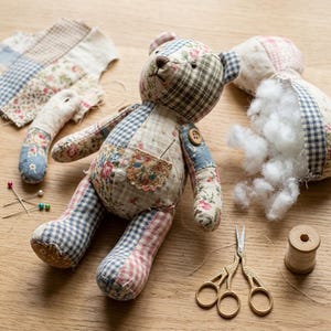 May include: A patchwork teddy bear made from various floral and gingham fabric scraps, featuring a small pocket, a button joint, and a needle. Nearby are sewing supplies including scissors, a wooden spool of thread, and straight pins.