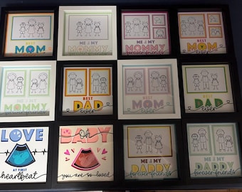 May include: A collection of twelve square shadow box frames featuring stick figure family illustrations, ultrasound images, and text such as Best Mom, Best Dad, Love at First Heartbeat, and Oh Baby, with colorful borders and black frames.