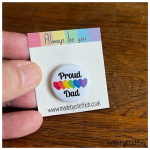May include: White button with the text "Proud Dad" in black lettering.  The button has a rainbow of hearts around the text.  The button is on a white card with the text "Always be you." and a website address.
