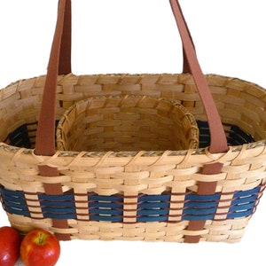 Puede incluir: Una cesta ovalada tejida con dos asas de cuero marrón. La cesta tiene una cesta tejida más pequeña en su interior y una banda de patrón azul y marrón alrededor del borde. Dos manzanas rojas están delante de la cesta.