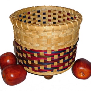 May include: A woven basket with a brown, red, and blue checkered pattern. The basket has a wooden base with three legs and is surrounded by three red apples.